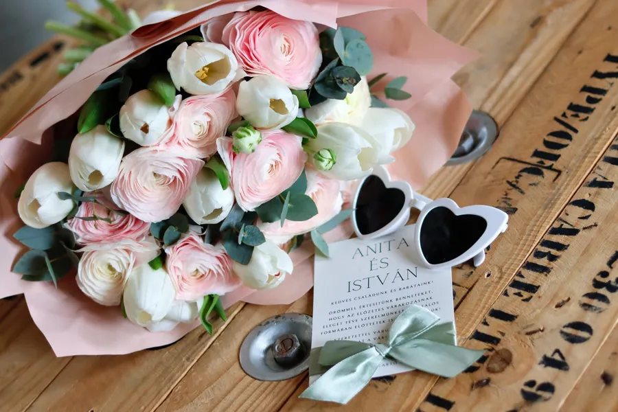 Esküvői flatlay: rózsaszín boglárka csokor, meghívó és szív alakú napszemüveg fa asztalon — Gáspár Enikő Photography